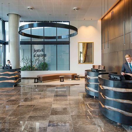 풀먼 브리즈번 공항 호텔 외부 사진 The image shows a modern hotel lobby. There are two reception desks made of dark-colored materials, and staff members are attending to guests at these desks. The lobby features large windows that allow natural light to fill the space, and there are p