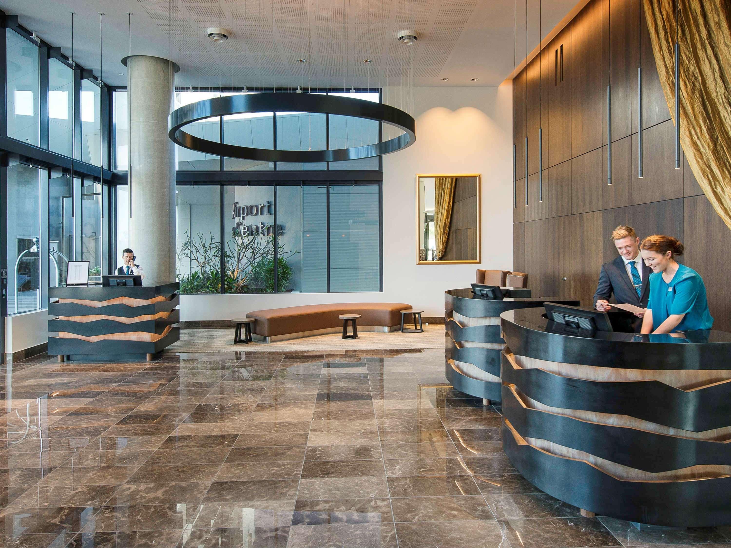 풀먼 브리즈번 공항 호텔 외부 사진 The image shows a modern hotel lobby. There are two reception desks made of dark-colored materials, and staff members are attending to guests at these desks. The lobby features large windows that allow natural light to fill the space, and there are p